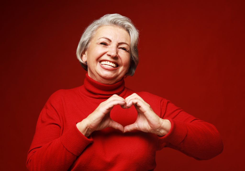 enior grey-haired woman wearing red sweater smiling in love doing heart symbol shape with hands.